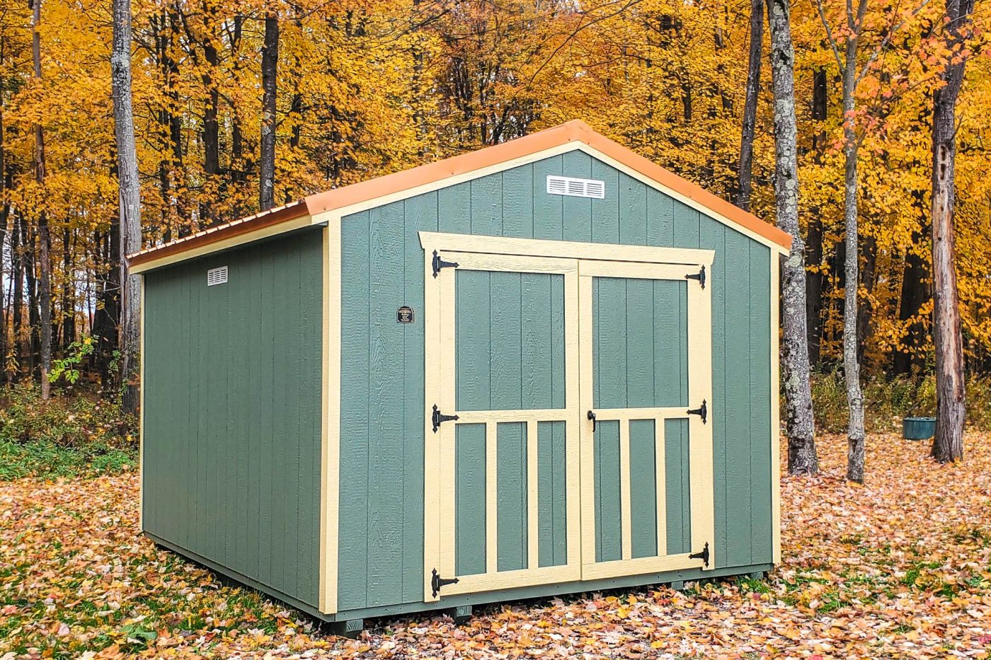 10x16 green utility shed with tan trim and barn doors, surrounded by autumn trees and fallen leaves.